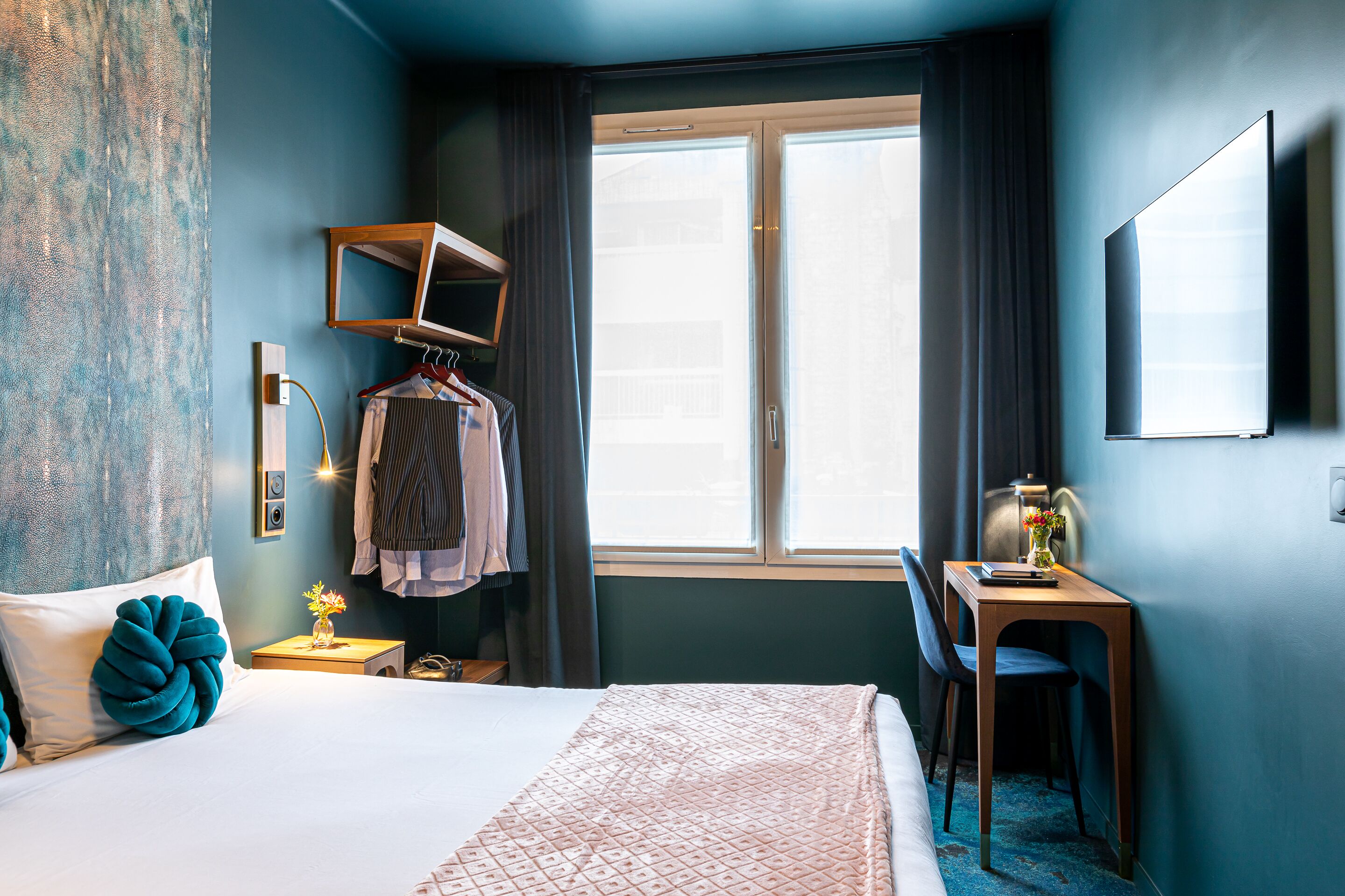 Une chambre d’hôtel comprenant un lit, un bureau avec une chaise, un téléviseur fixé au mur et une fenêtre avec des rideaux.