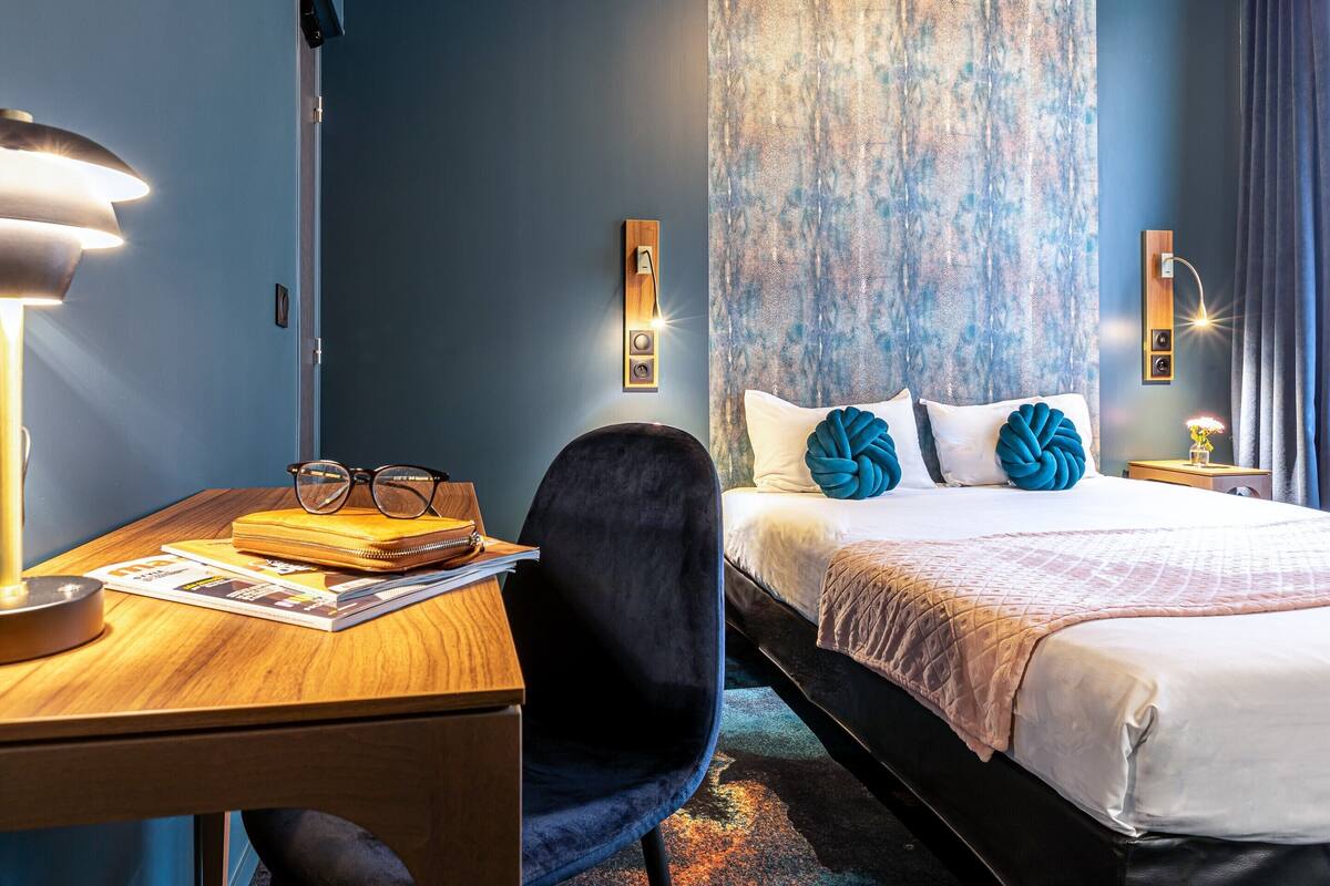 Une chambre d’hôtel avec un bureau en bois, une chaise noire, un lit recouvert d’une courtepointe blanche et rose, et un mur bleu à la texture particulière.