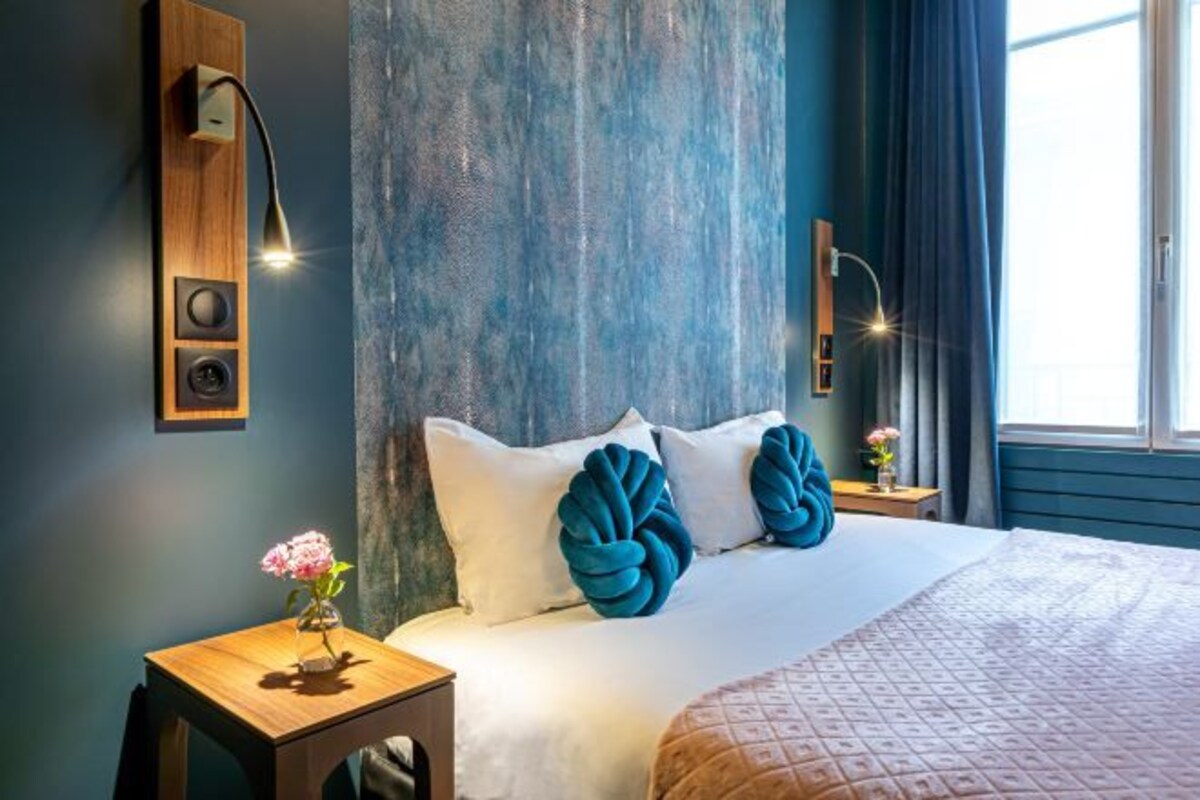 Ett modernt hotellrum med en stor säng, ett träbord vid sängen med en vas med blommor och ett stort fönster med gardiner.