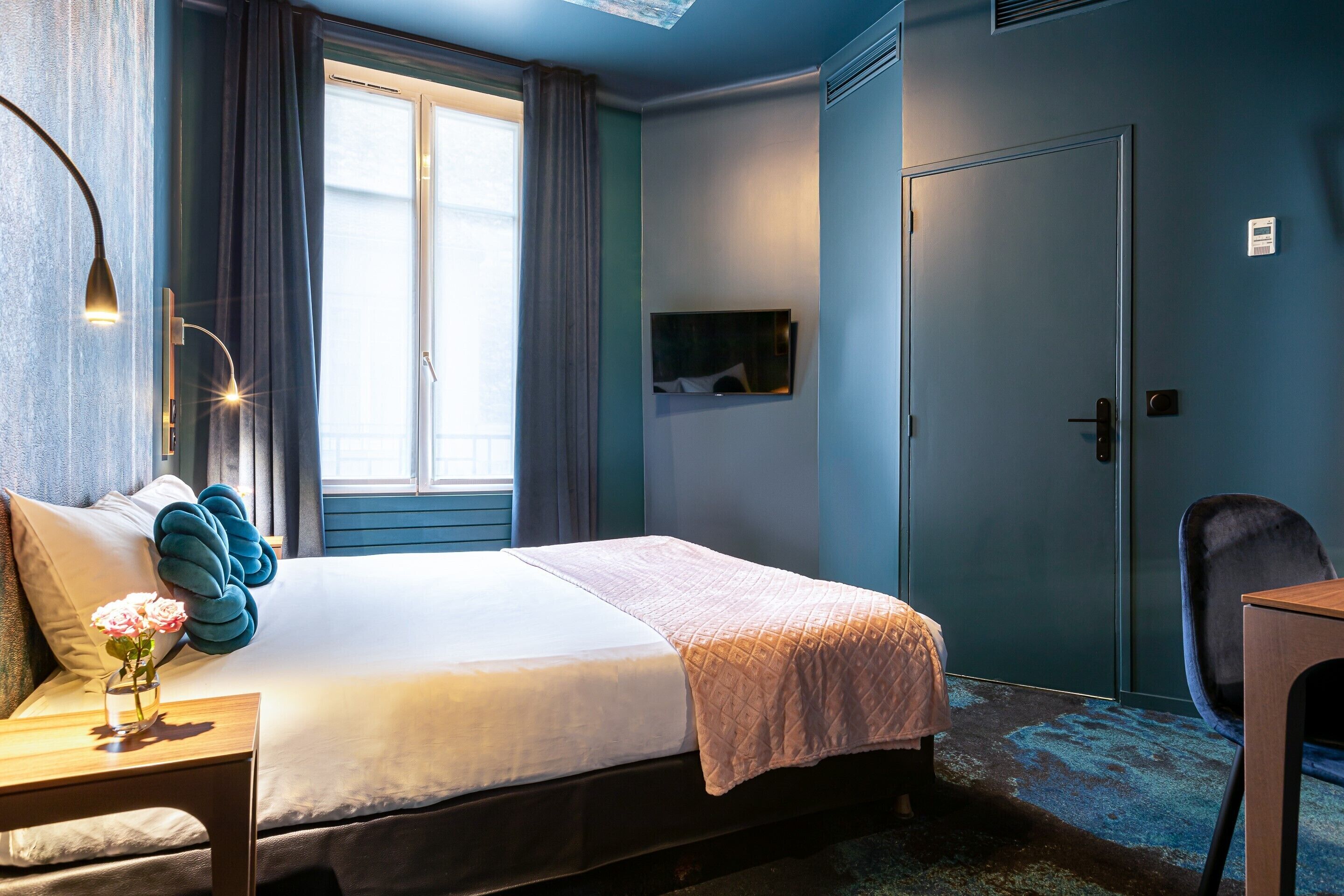 Quarto de hotel com cama, escrivaninha com luminária, cadeira, televisão e porta.