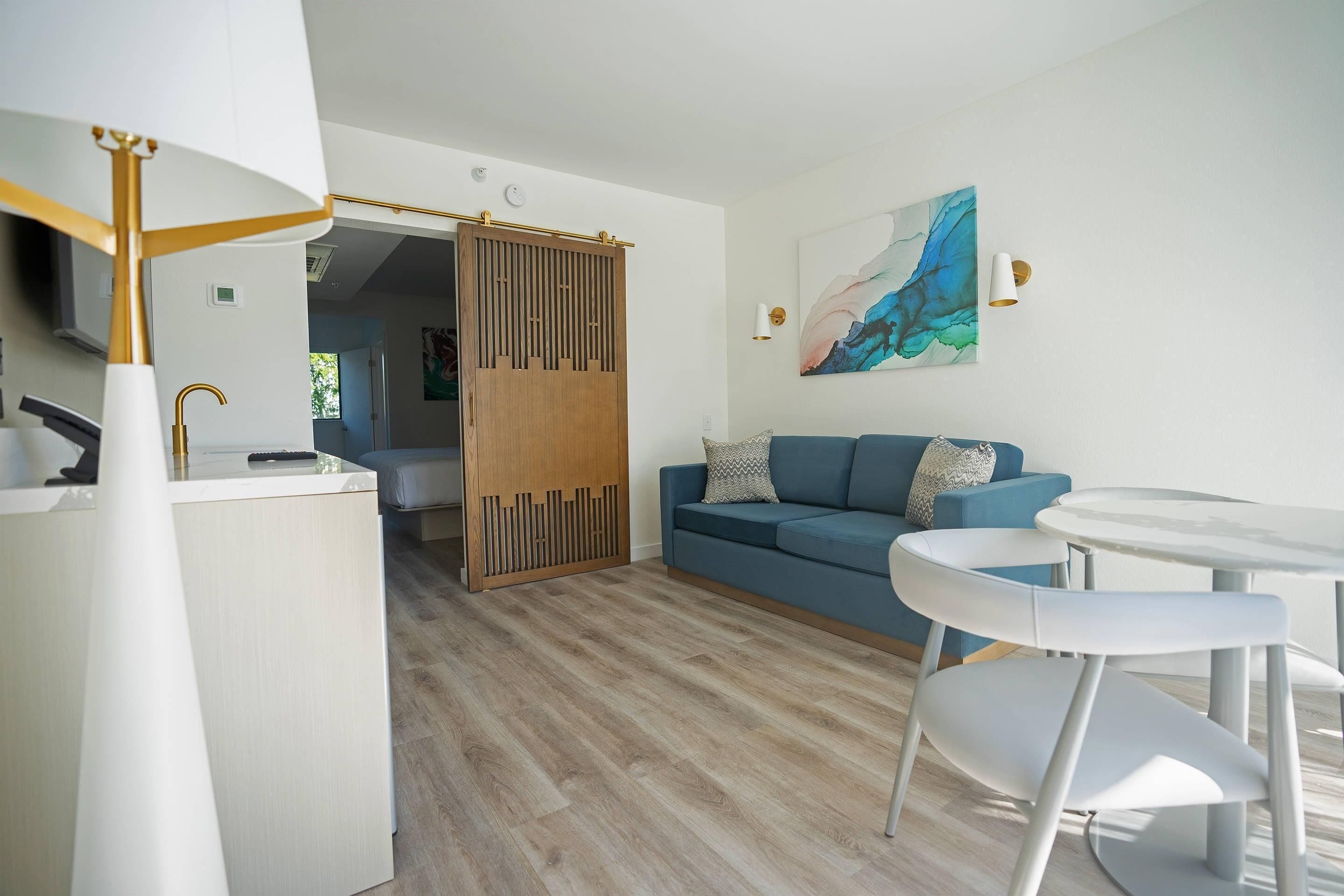 Una sala de estar moderna con un sofá azul, sillas blancas, una mesa de vidrio, un separador de madera y un cuadro en la pared.