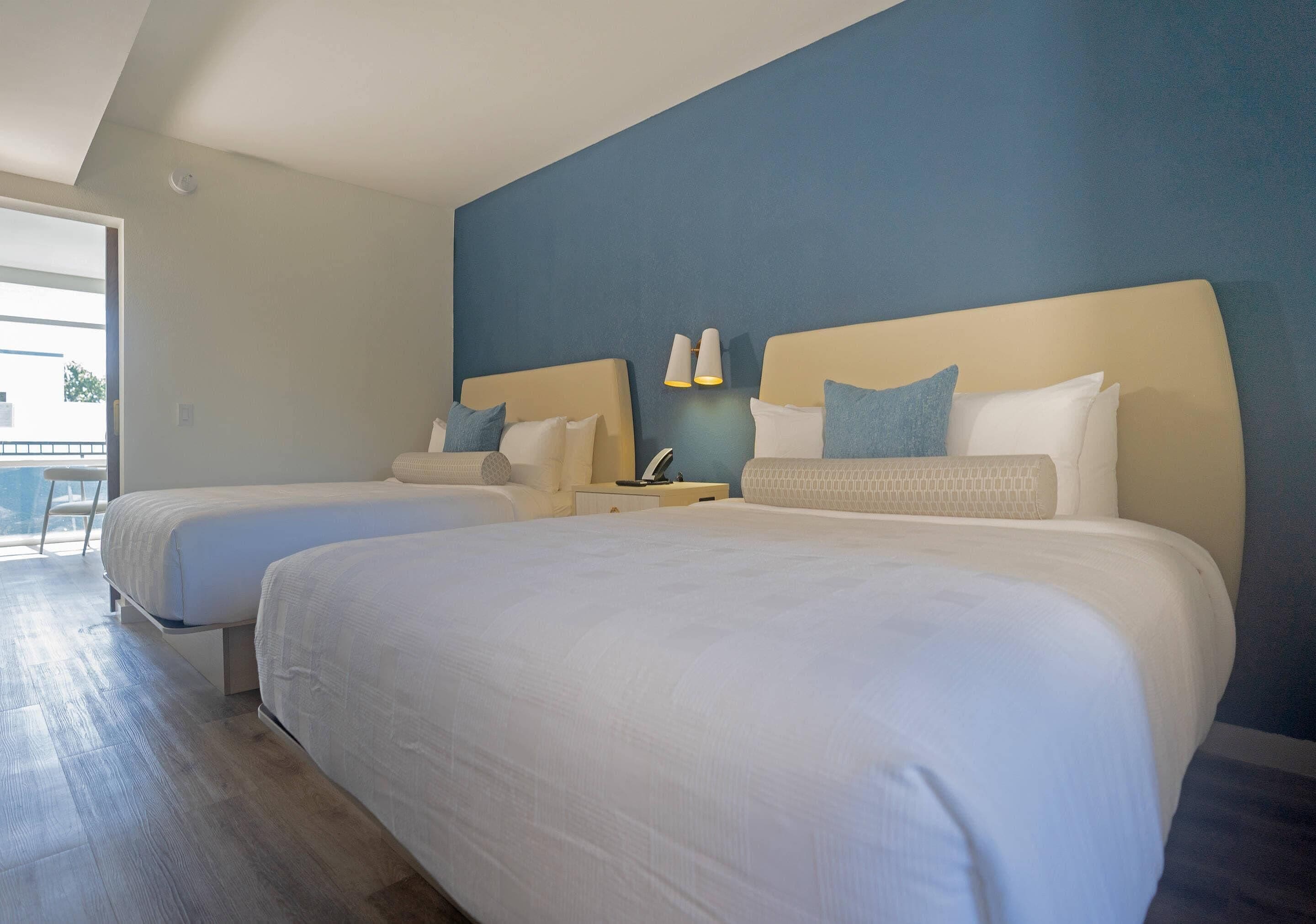Habitación de hotel con dos camas, una mesita de noche, una lámpara y vistas al exterior.