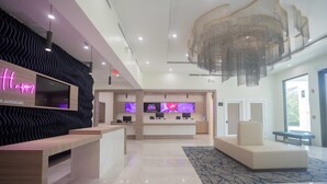 A modern hotel lobby with a reception desk, multiple TVs, a chandelier, and a seating area.