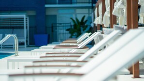 A row of sun loungers by a poolside, with a blue building and a plant in the background.