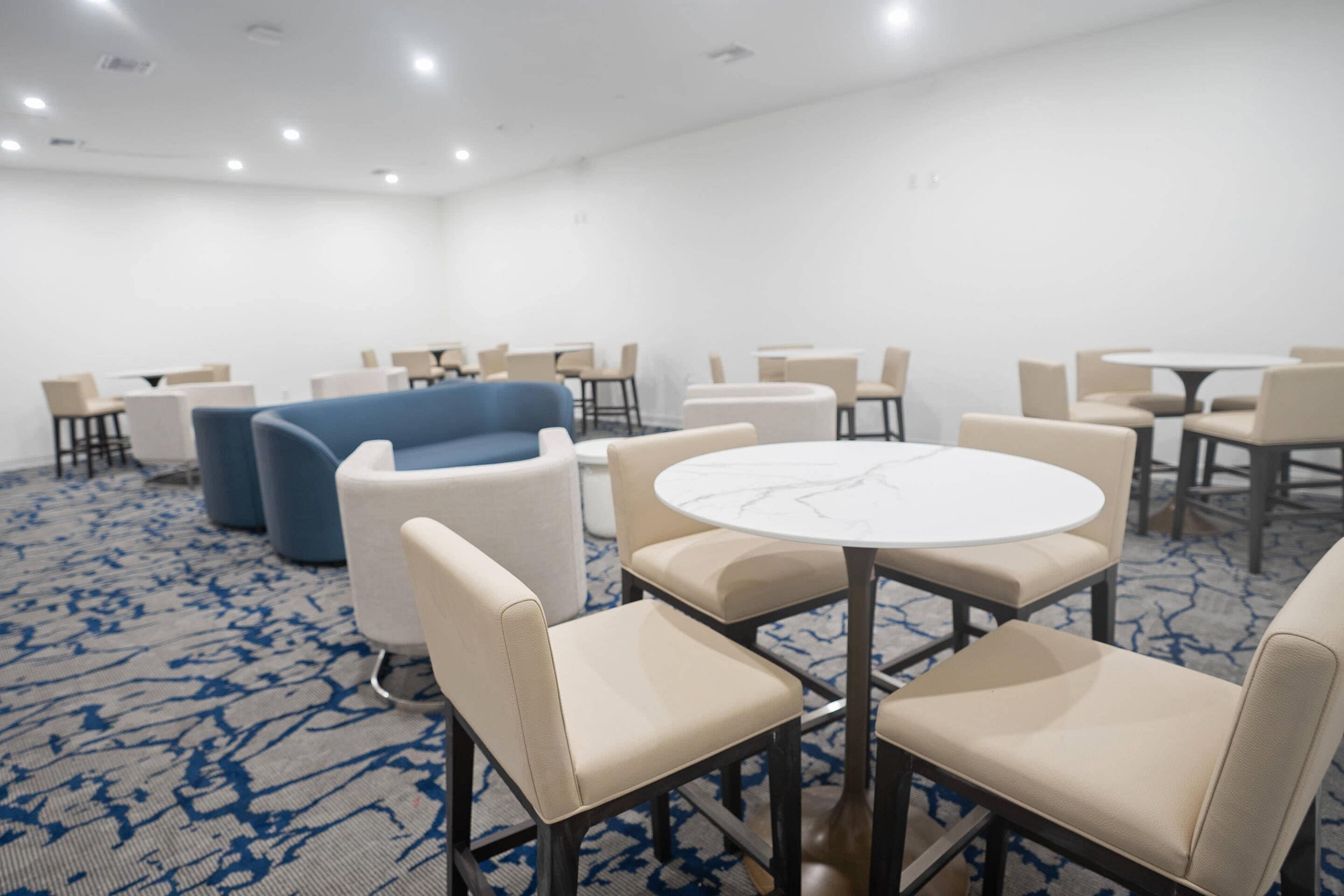 Une salle de conférence avec des tables rondes et des chaises beiges, un canapé bleu et un sol recouvert de moquette.