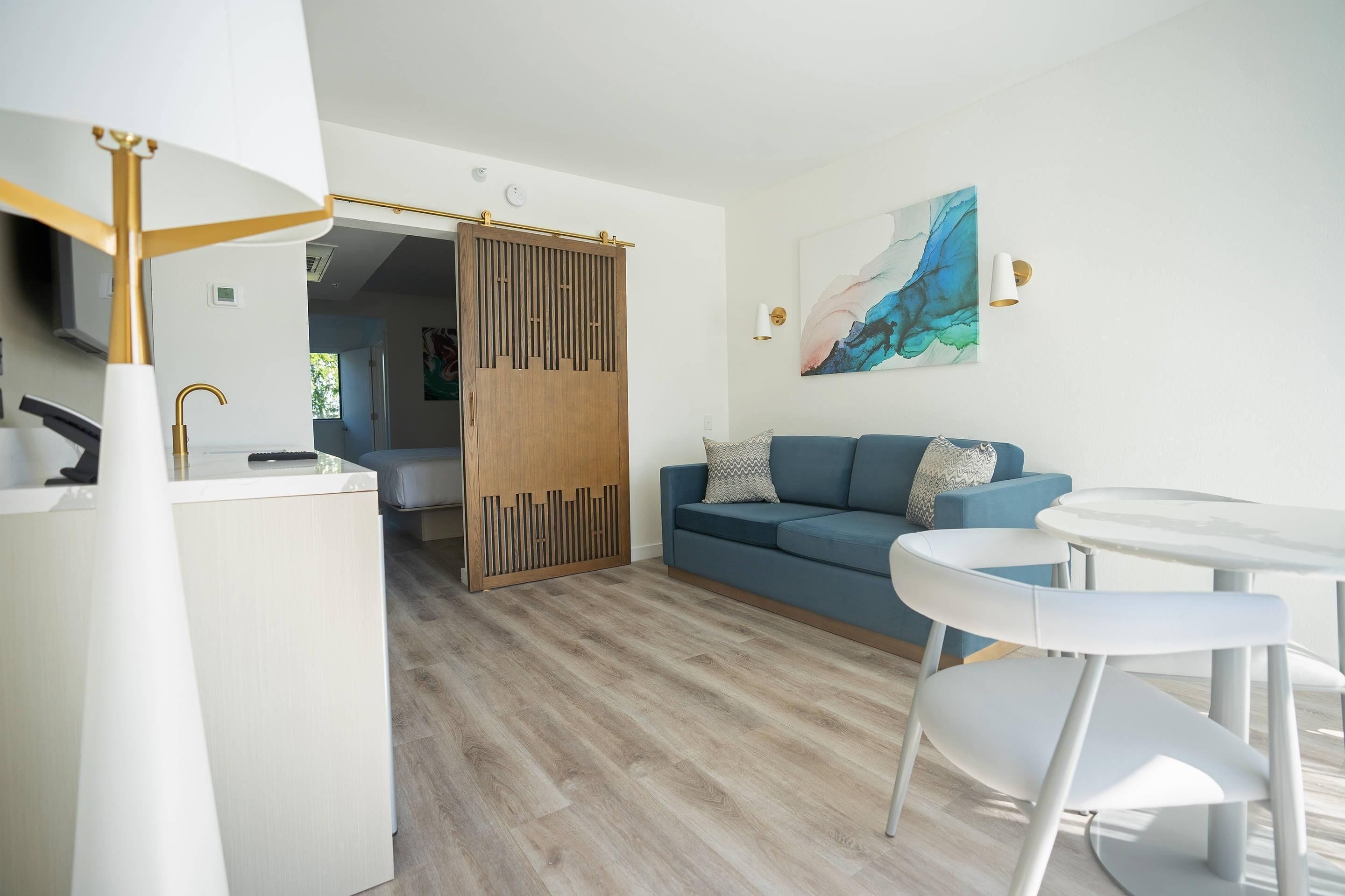 Een moderne woonkamer met een blauwe bank, witte stoelen, een glazen salontafel en een houten roomdivider.