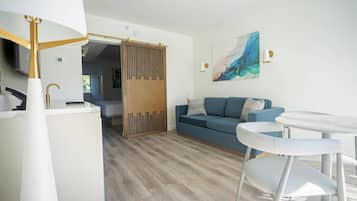 Een moderne woonkamer met een blauwe bank, witte stoelen, een glazen salontafel en een houten roomdivider.