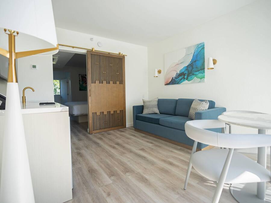 Een moderne woonkamer met een blauwe bank, witte stoelen, een glazen salontafel en een houten roomdivider.