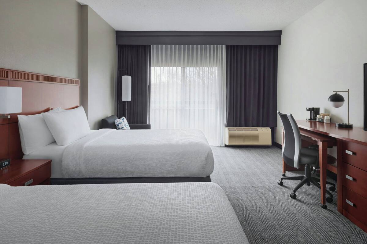 A hotel room with two beds, a desk, a chair, a lamp, and a large window with curtains.