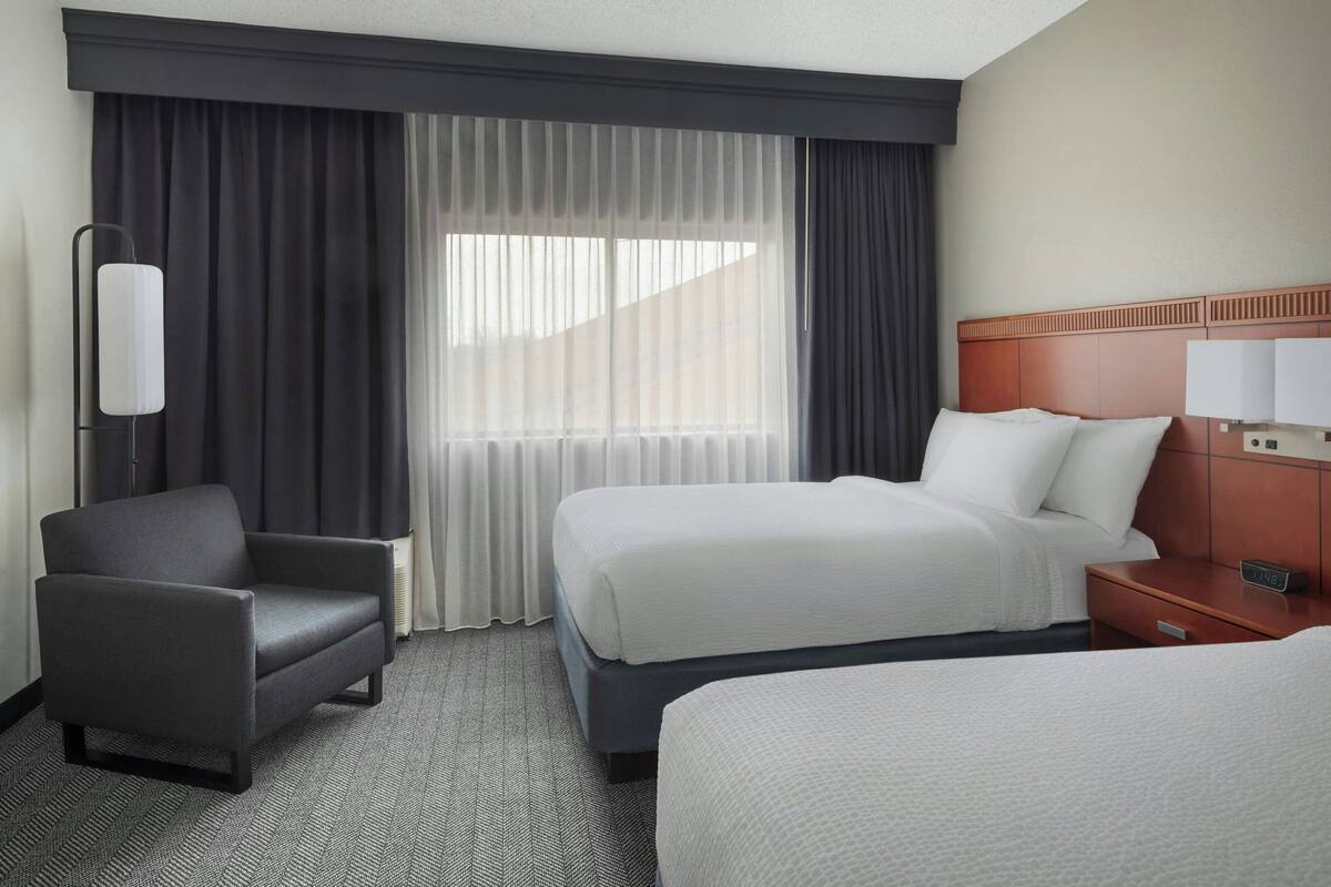 A hotel room with a bed, a chair, a nightstand, and a window with curtains.