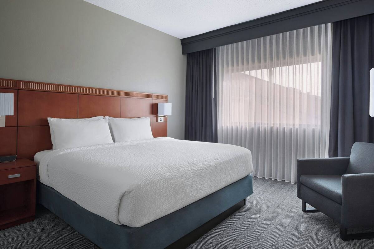 A hotel room with a large bed, a nightstand, a chair, and a window with curtains.