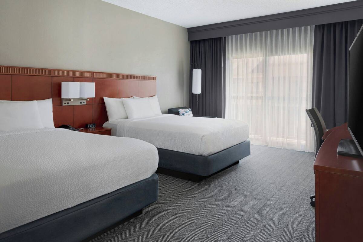 A hotel room with two beds, a desk, a lamp, and a large window with curtains.