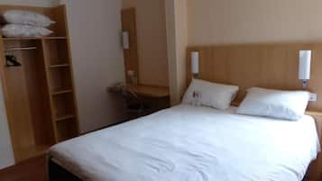 A hotel room with a bed, a wooden wardrobe, a desk with a chair, and a small trash bin.