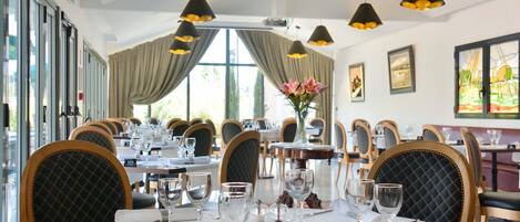 A dining area with tables set for six, each with a white tablecloth, wine glasses, and cutlery. There are black chairs with a diamond pattern on the backrests. The room has large windows with curtains, and pendant lights hang from the ceiling.