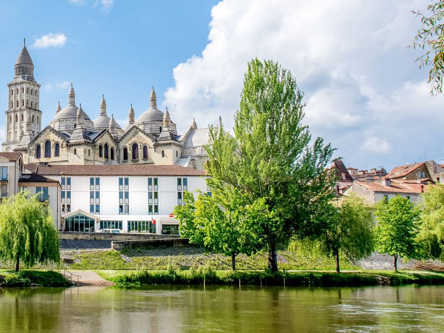 Un edificio histórico con múltiples cúpulas y una torre, ubicado junto a un río tranquilo y abundante vegetación.