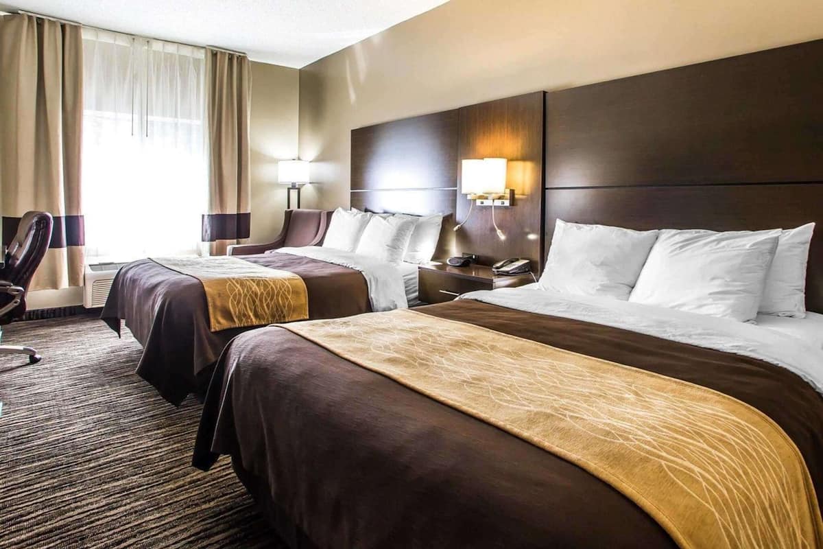 A hotel room with two beds, a desk, a chair, and a lamp.