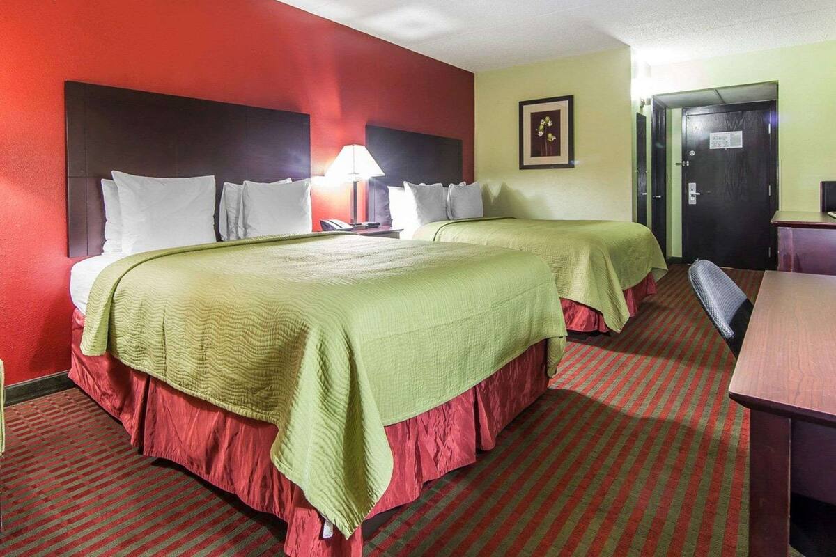 A hotel room with two beds, a desk, a chair, a lamp, and a door.