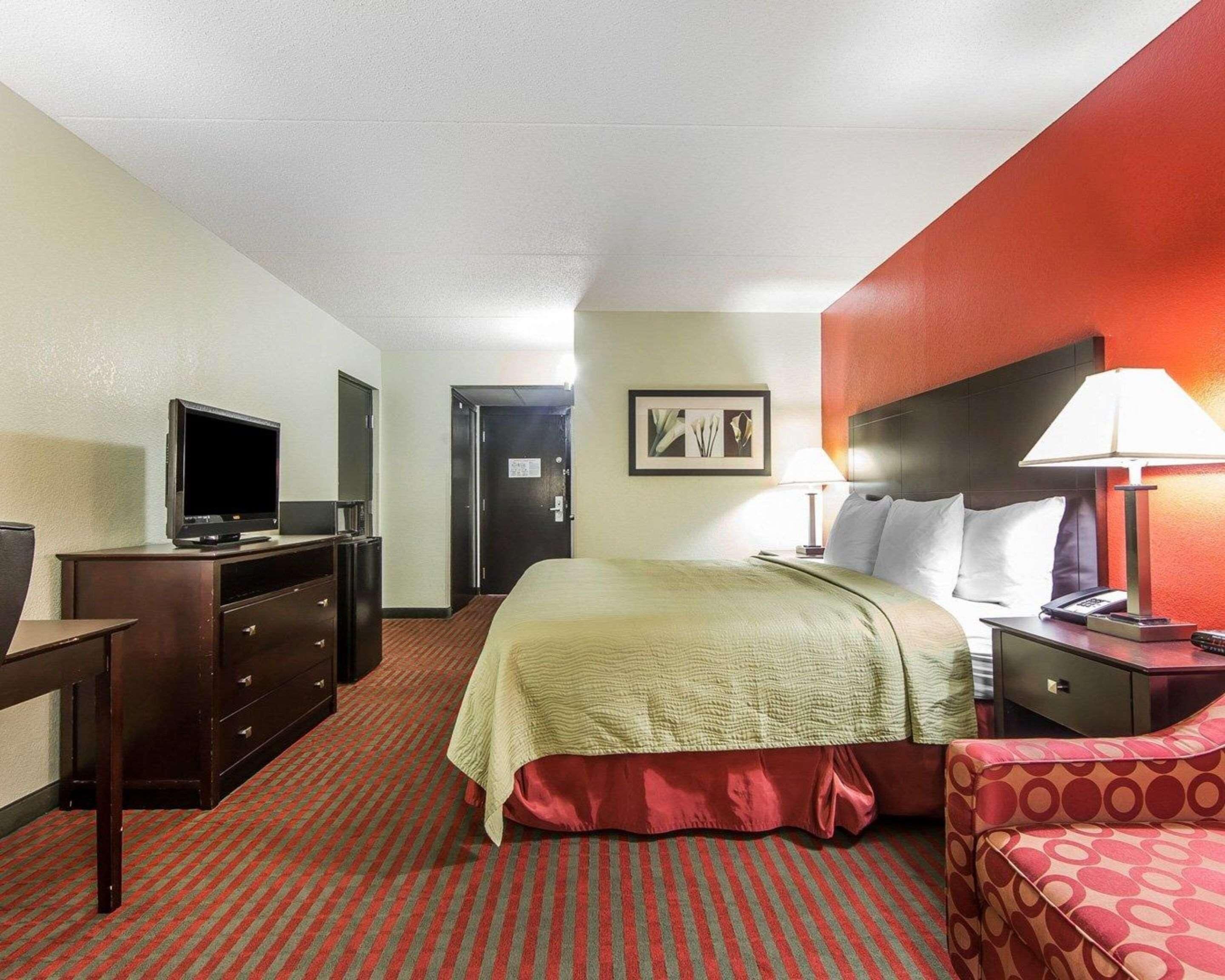 Habitación de hotel con cama, escritorio con televisor, cómoda, mesita de noche con lámpara y puerta que da a otra habitación.