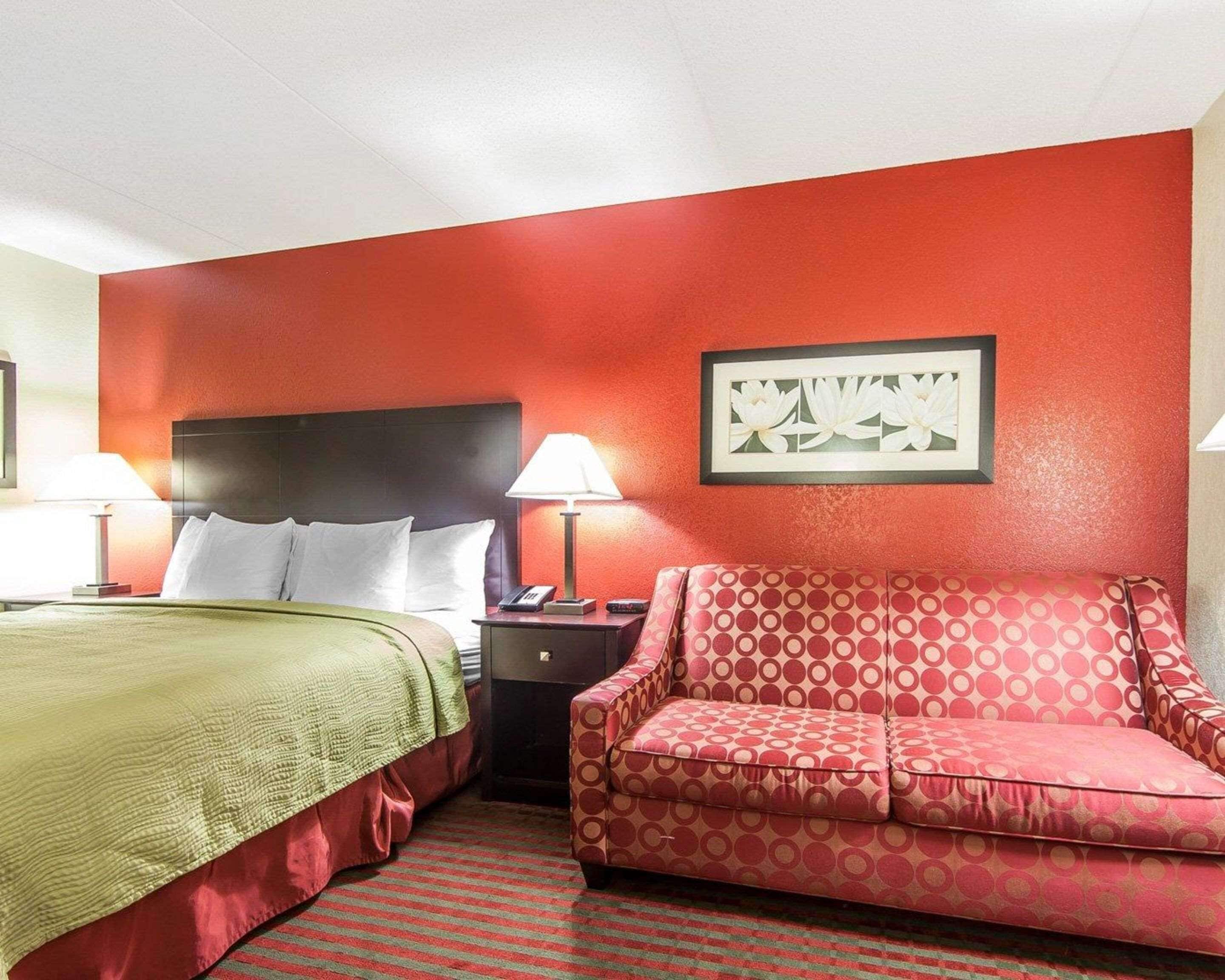 Habitación de hotel con una cama, un sofá rojo, una mesita de noche con lámpara y un cuadro en la pared.