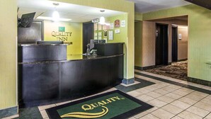 A hotel reception area with a reception desk, a check-in counter, and a reception desk with a computer. There are signs on the wall and a green carpet with the hotel's logo. - Quality Inn Sweetwater West (Sweetwater)