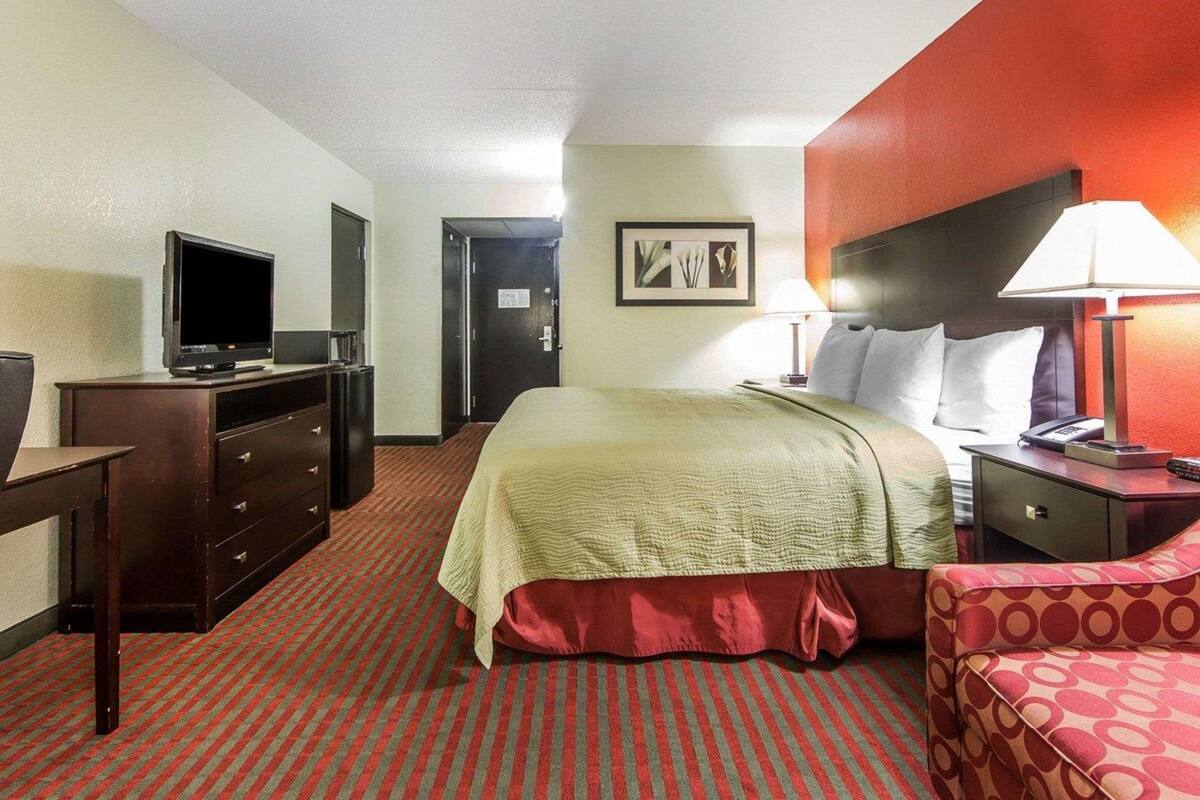 A hotel room with a bed, a desk with a television, a dresser, a nightstand with a lamp, and a door leading to another room.