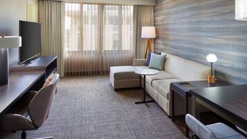 Quarto de hotel com televisão de tela plana, sofá, mesa redonda pequena, escrivaninha e luminária.