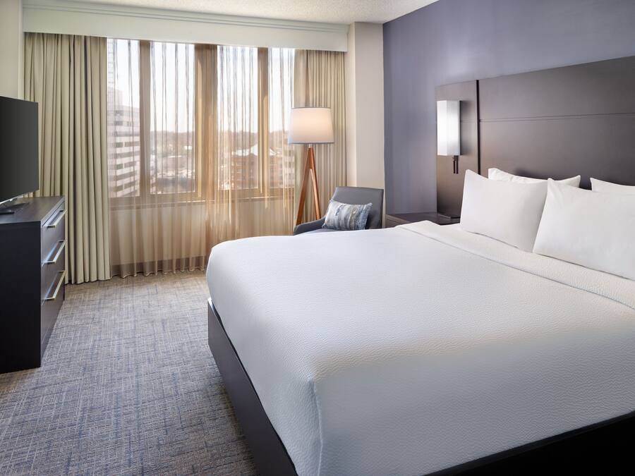 Quarto de hotel com uma cama grande, televisão de tela plana, escrivaninha com luminária e vista da cidade através de cortinas finas.