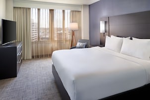 Habitación de hotel con una cama grande, televisor de pantalla plana, escritorio con lámpara y vistas a la ciudad a través de cortinas traslúcidas.