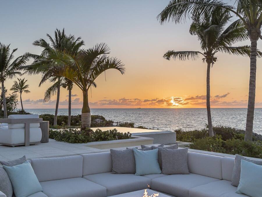 Un área de estar moderna al aire libre con un elemento decorativo de fuego, rodeada de palmeras y con vistas al océano al atardecer.