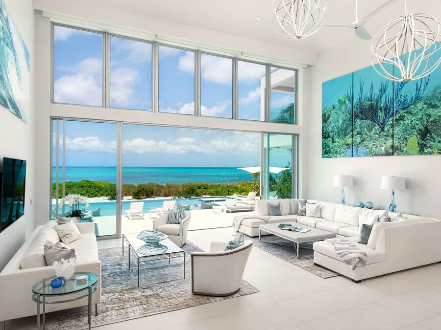 Amplia sala de estar con un gran ventanal que da a una playa y al océano, muebles blancos y una lámpara moderna.