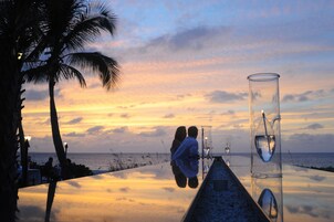 Una mesa junto a la playa con un vaso de agua y una vela, palmeras y vista al atardecer.
