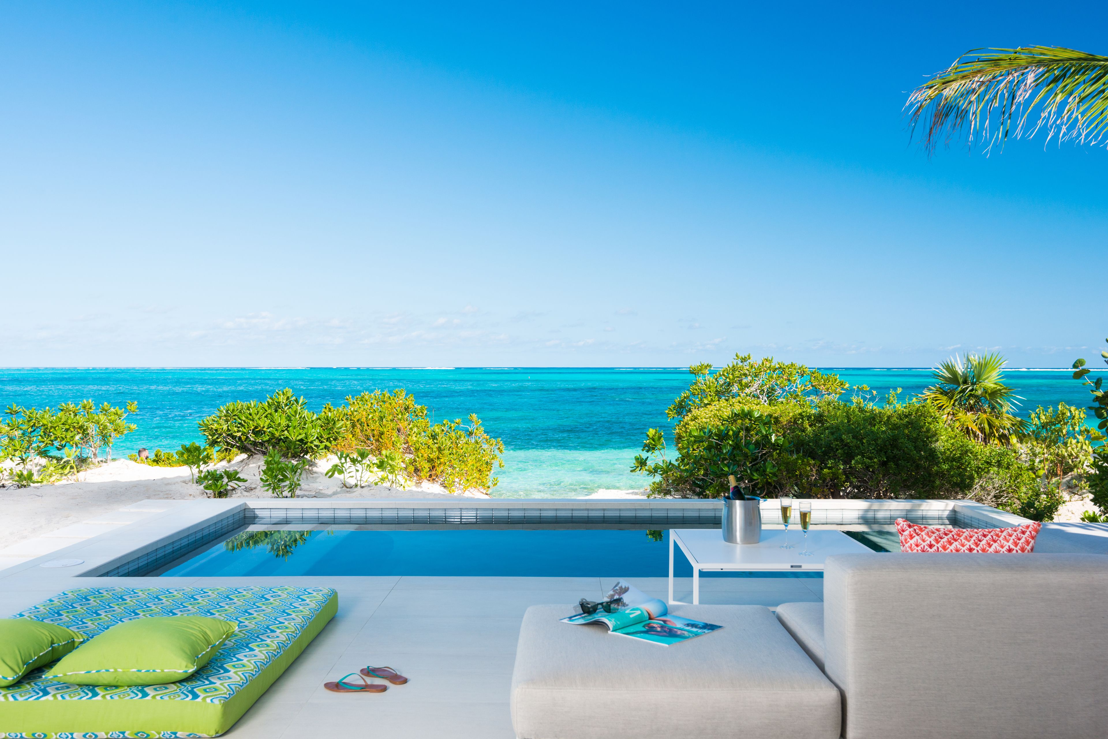 A poolside area with a view of the ocean, a lounge with a sofa and cushions, and a small table with a drink.