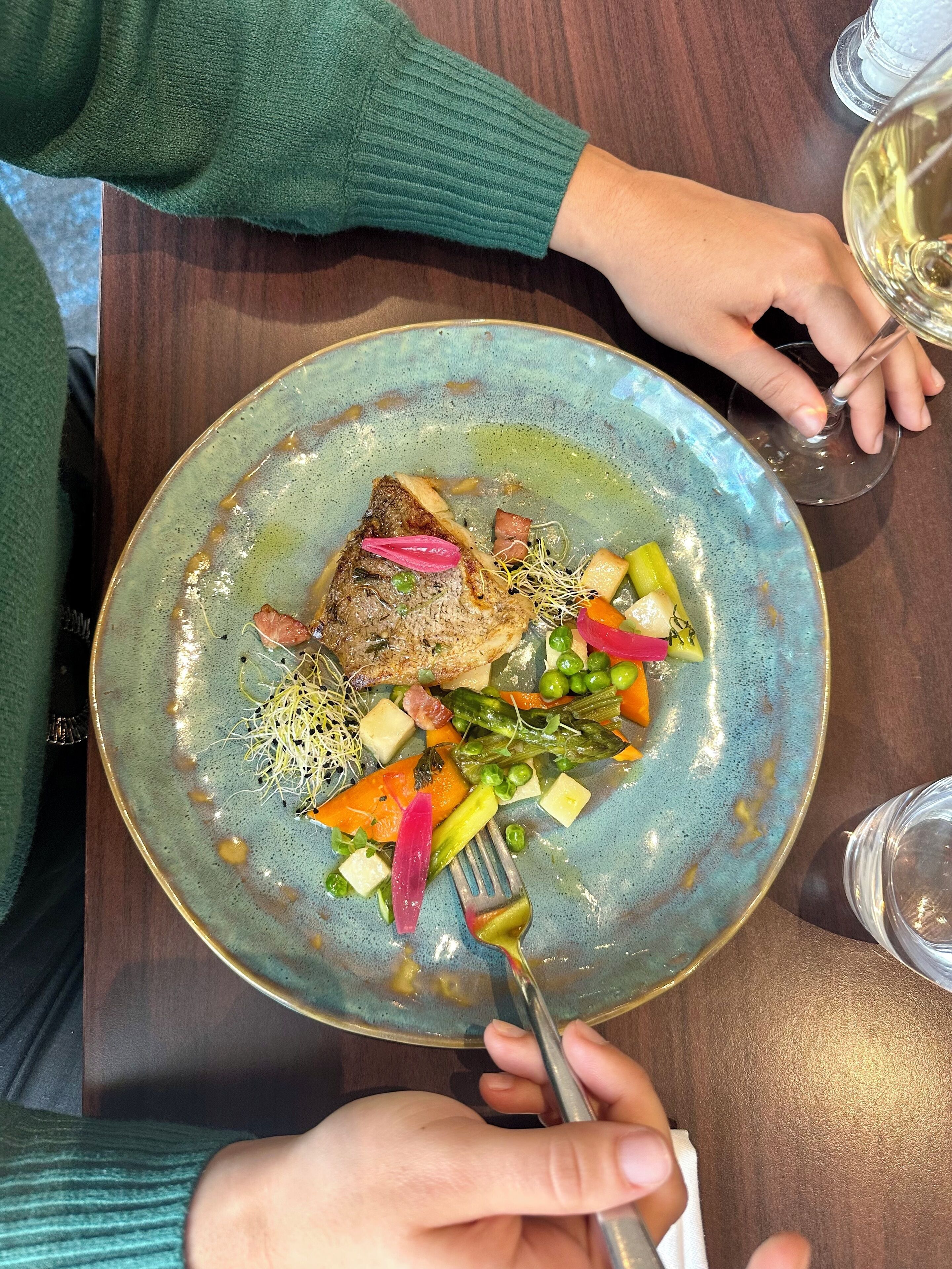 Une personne tenant une fourchette devant une assiette remplie de nourriture, notamment du poisson et des légumes, sur une table.