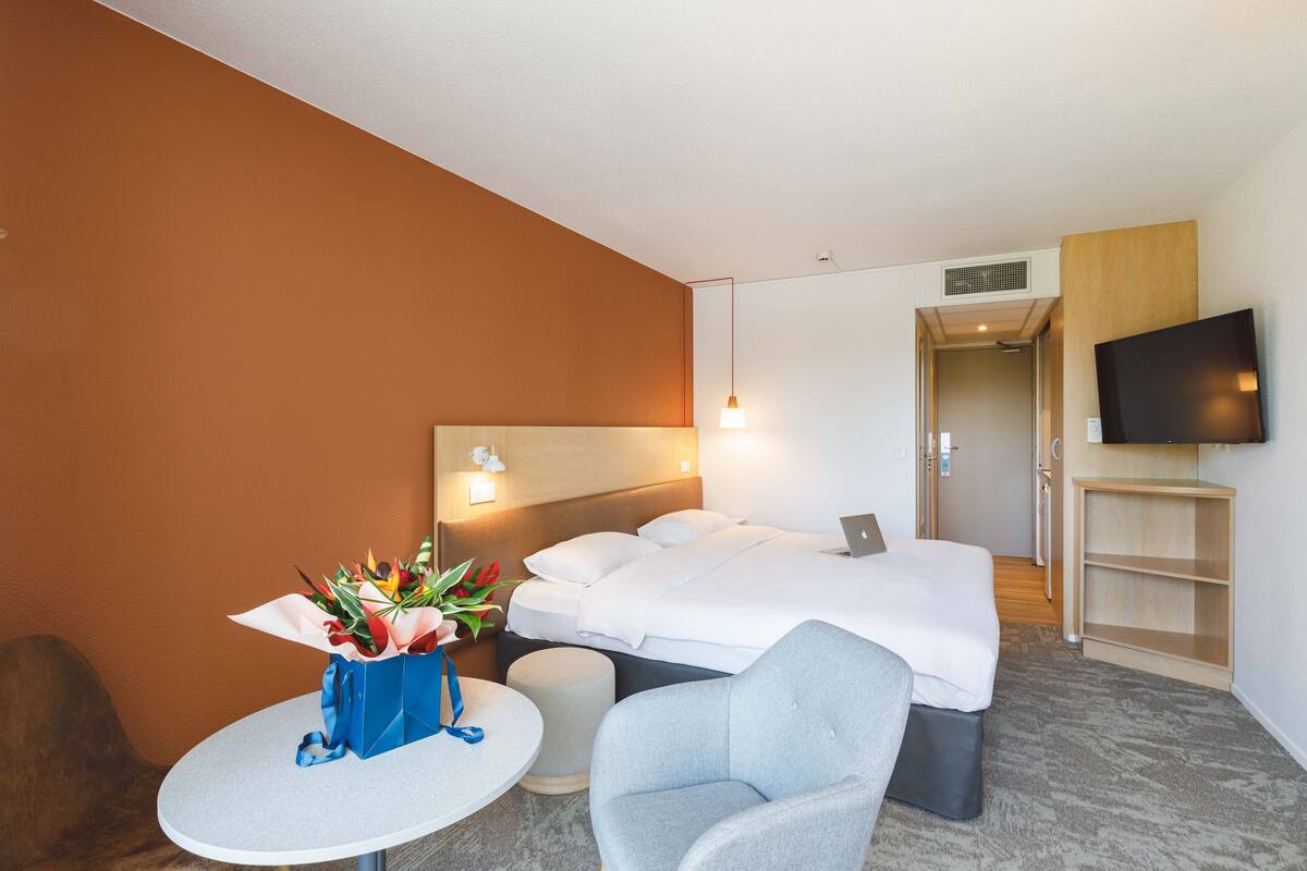 Une chambre d’hôtel avec un lit, un bureau avec un ordinateur portable, un téléviseur fixé au mur et une petite table sur laquelle est posé un vase de fleurs.
