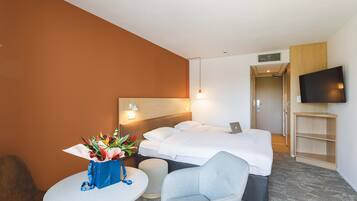 Une chambre d’hôtel avec un lit, un bureau avec un ordinateur portable, un téléviseur fixé au mur et une petite table sur laquelle est posé un vase de fleurs.