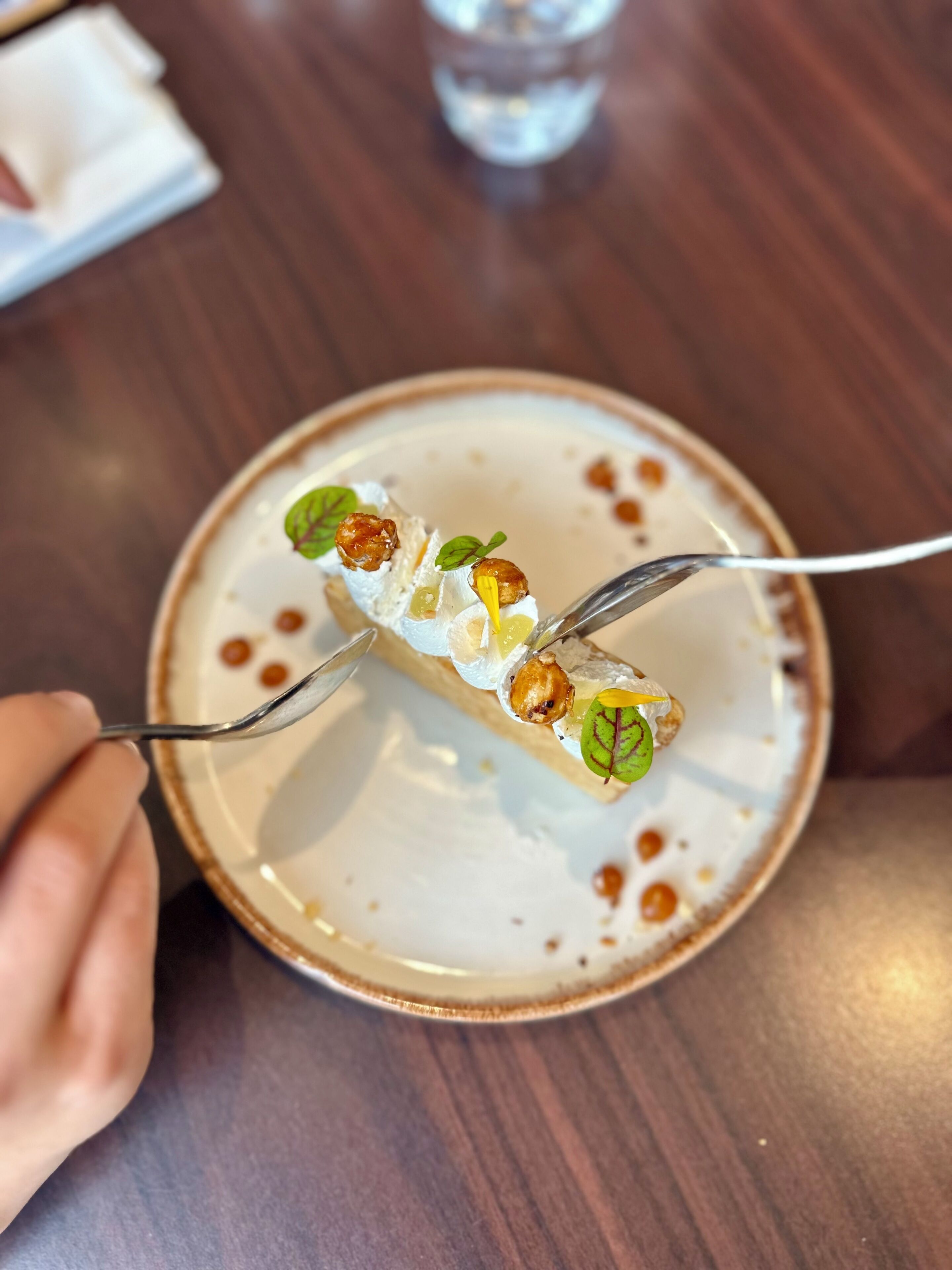 Une assiette contenant un dessert agrémenté de noix et de feuilles, une fourchette et un verre d’eau, le tout posé sur une table en bois.