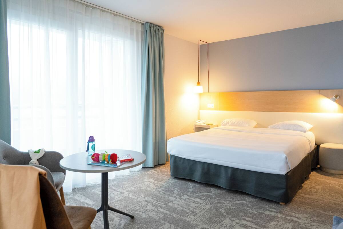 Une chambre d’hôtel avec un lit, une table ronde sur laquelle se trouvent une bouteille et un jouet, une chaise et une table de chevet avec une lampe.