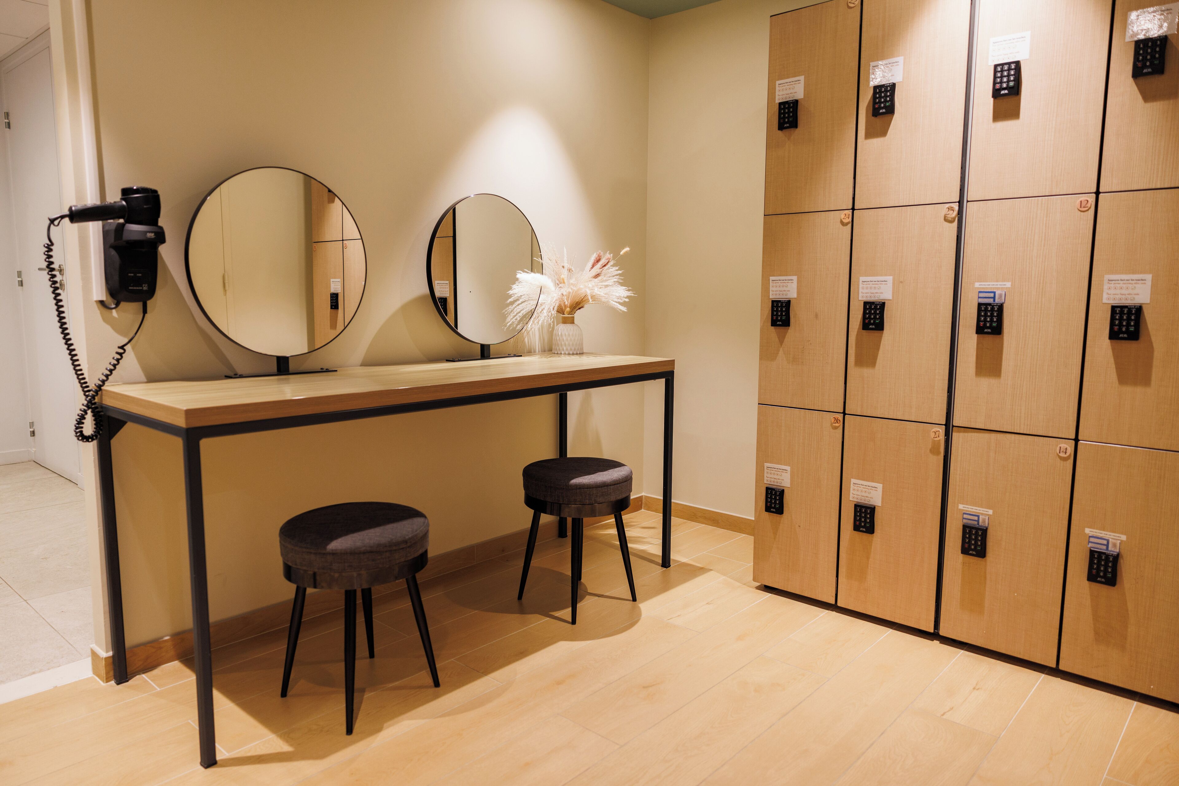 A room with a vanity table, two stools, a mirror, a hairdryer, and a row of lockers.