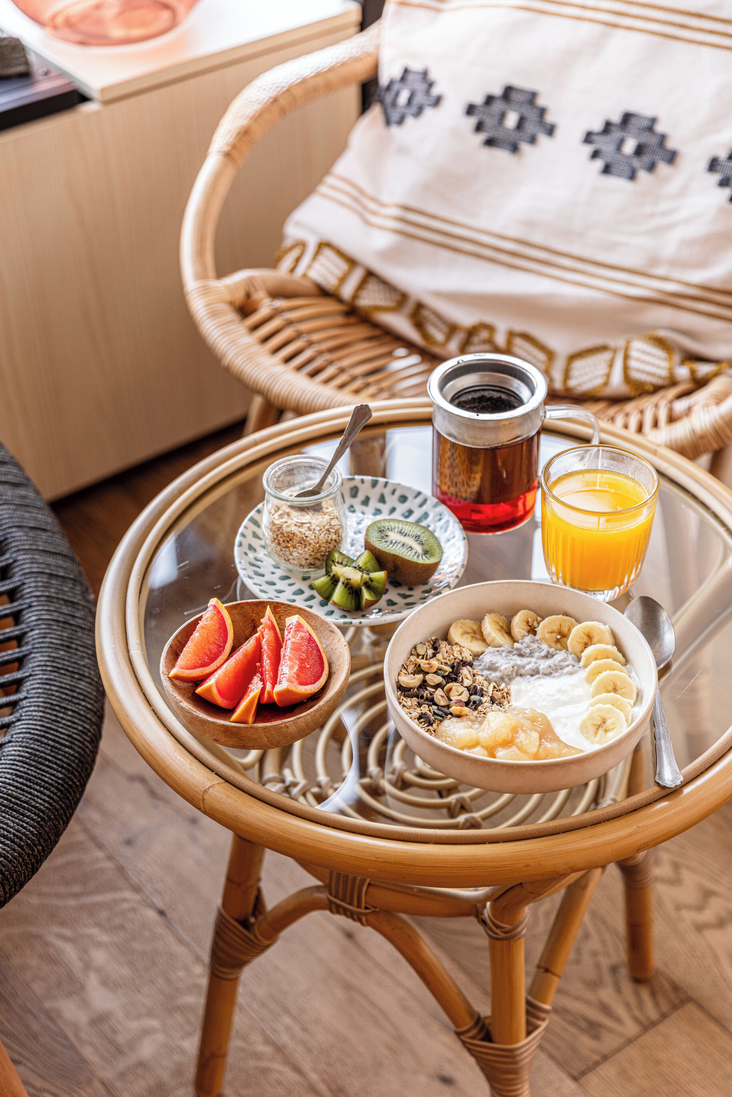 A wicker chair with a tray table holding breakfast items including a bowl of cereal, sliced bananas, grapefruit, a glass of orange juice, and a jar of honey.
