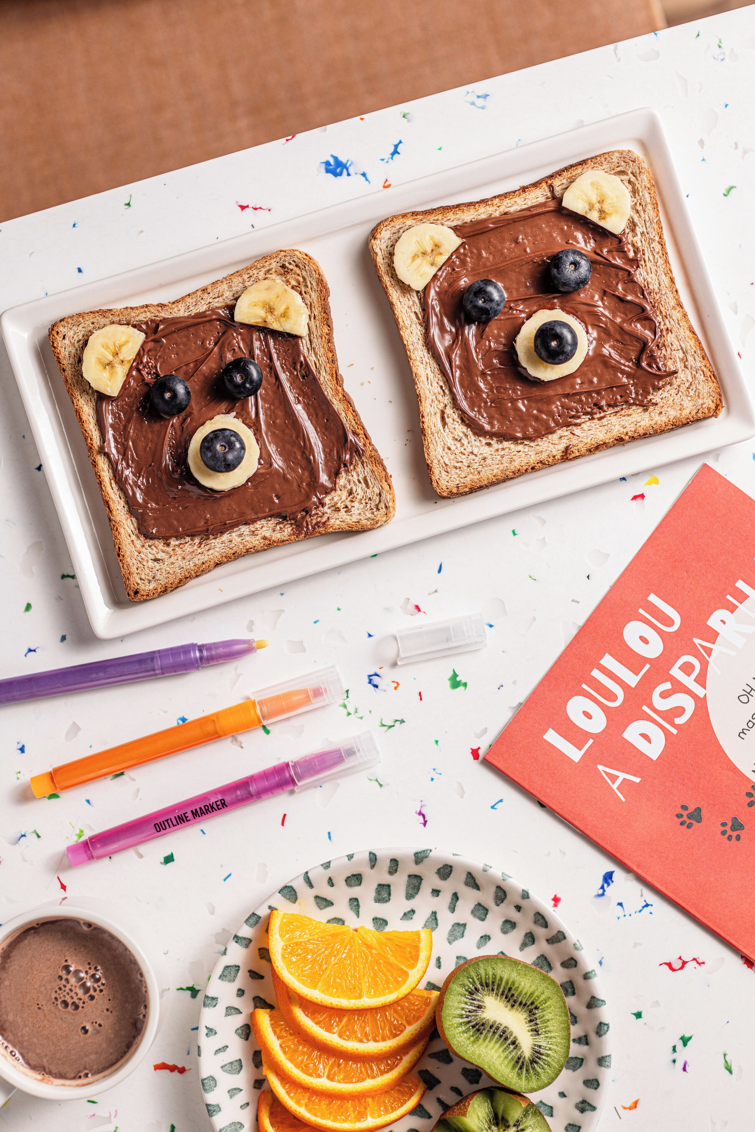 Zwei Scheiben Toast mit Schokoaufstrich, Bananen und Blaubeeren, die Gesichter bilden, ein Teller mit Orangenscheiben und Kiwis, eine Tasse Schokoladengetränk und ein rotes Buch mit dem Titel "LOU LOU A DISPARO".