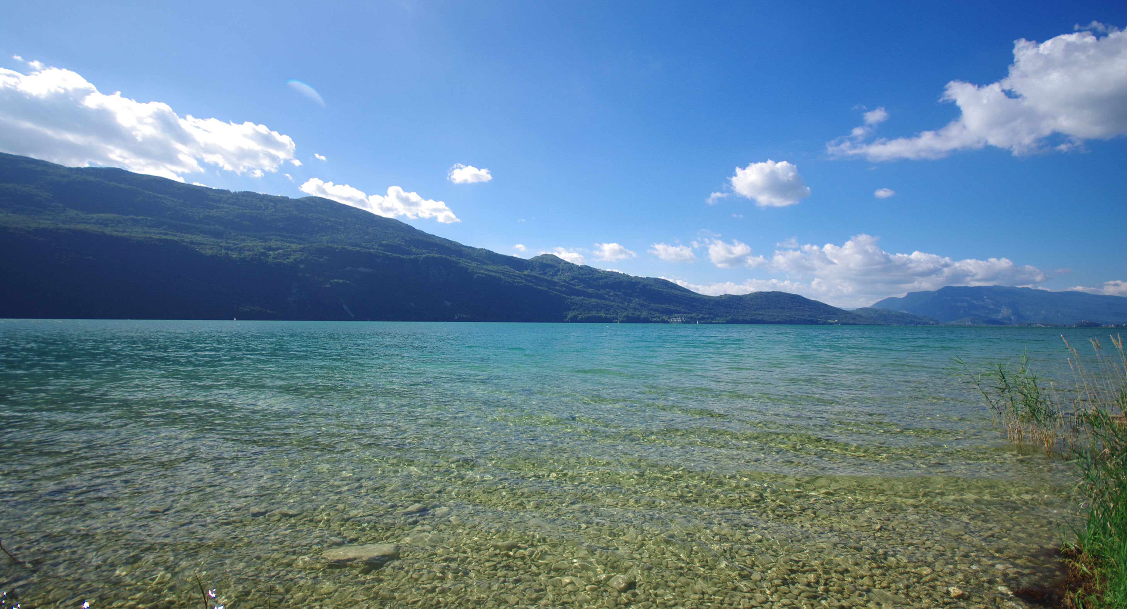 A clear lake with visible rocky bottom and distant mountains under a blue sky.