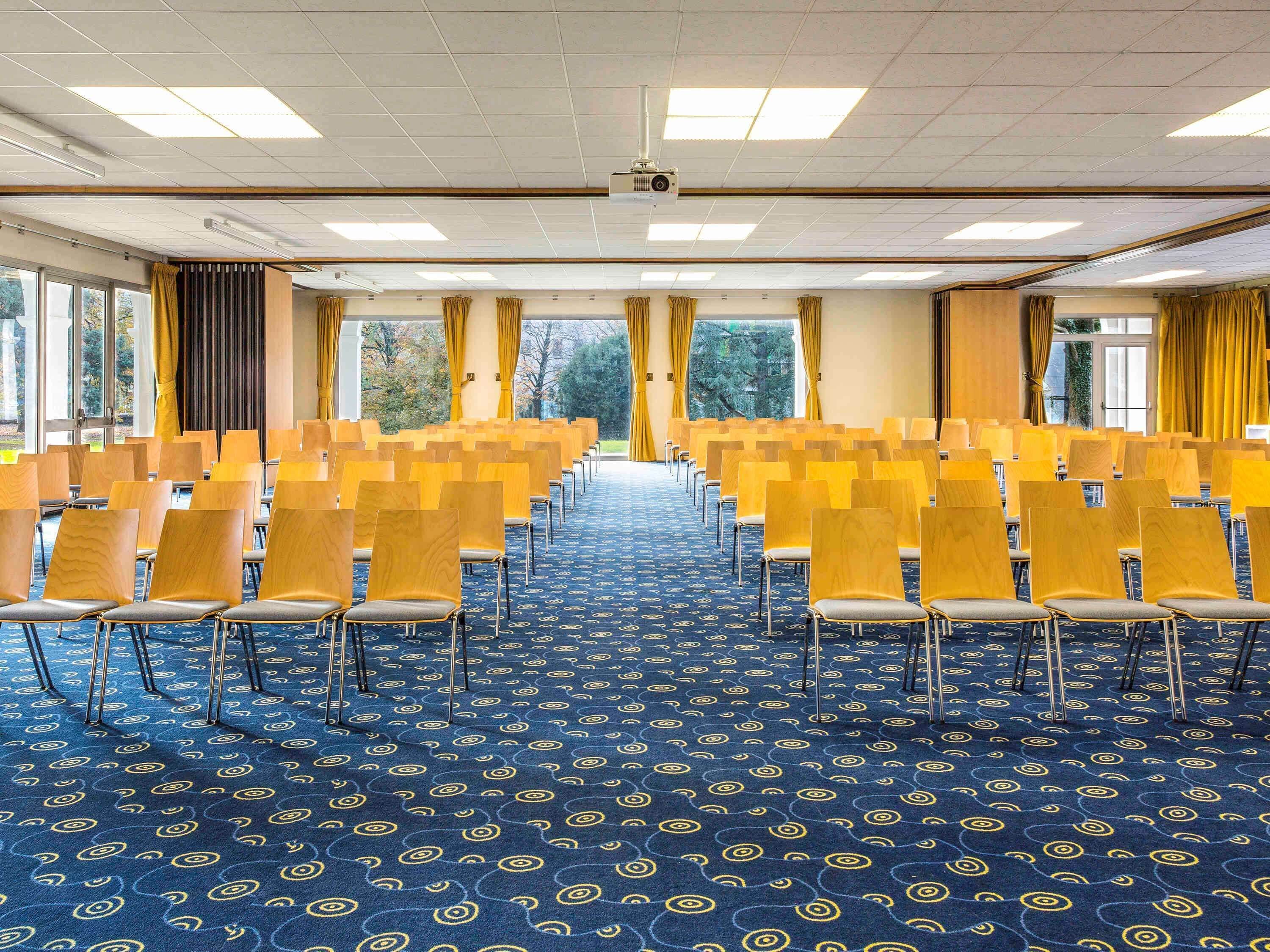 Un salón de conferencias con filas de sillas, una alfombra estampada y grandes ventanales con cortinas.