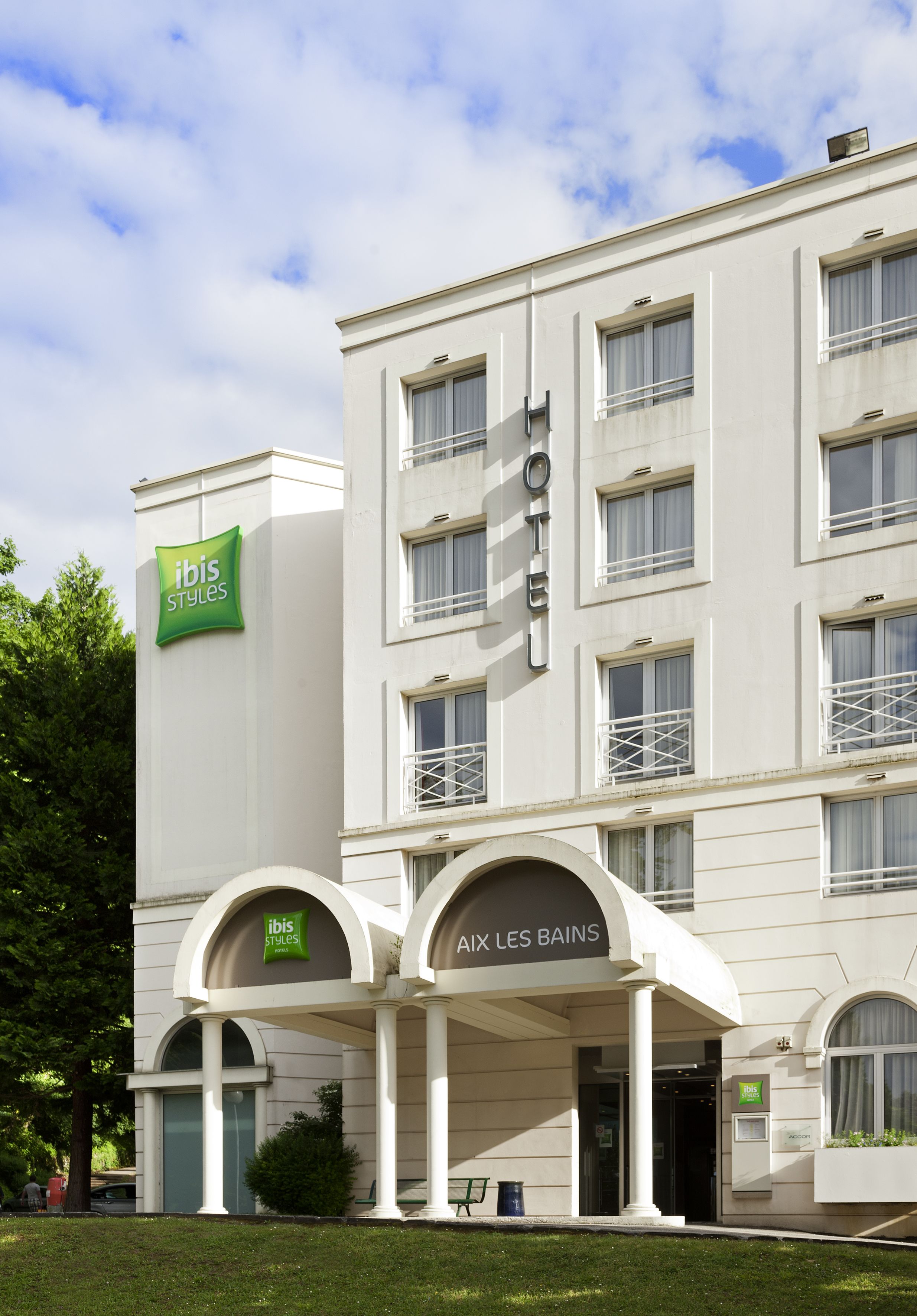 A hotel with a modern facade, featuring a prominent green 'ibis Styles' sign, a 'HOTEL' sign above the entrance, and a sign indicating 'AIX LES BAINS'.