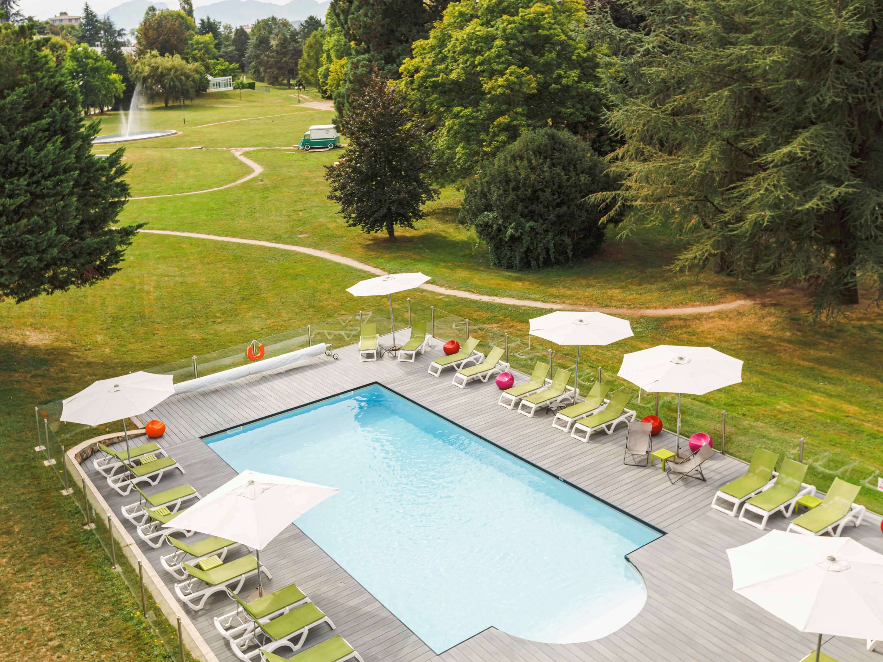 A swimming pool with lounge chairs and umbrellas, surrounded by a wooden deck and a grassy area with trees.