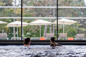 A hot tub area with two people, large windows, and outdoor seating with umbrellas.