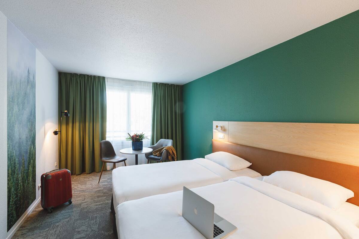Une chambre d’hôtel avec deux lits, un bureau avec un ordinateur portable, une chaise, une table avec une plante et une valise.