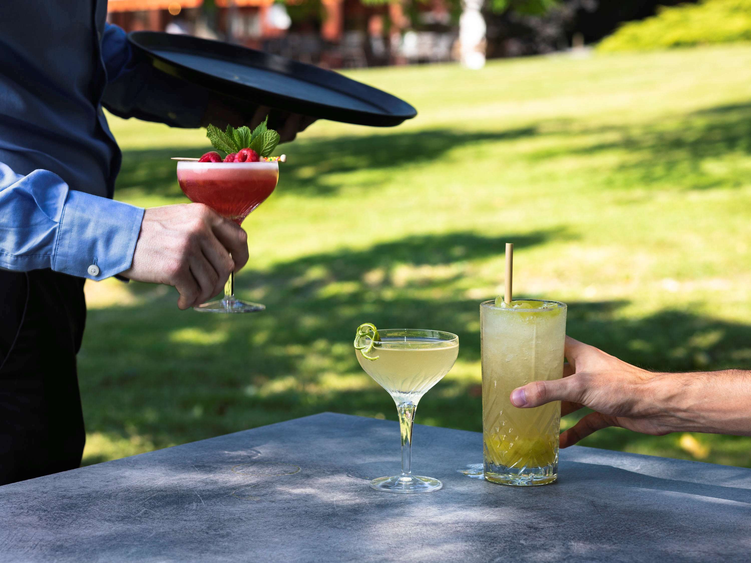 Eine Person hält ein Cocktailglas mit Strohhalm und Minze, daneben ein Glas mit Limette auf einem Tisch im Freien.