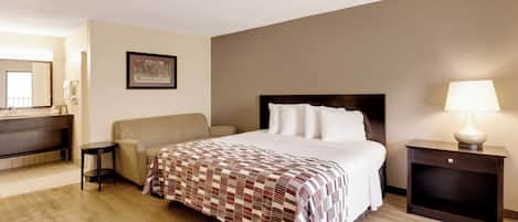 Habitación de hotel con una cama, una mesita de noche con lámpara, una silla, un sofá, un escritorio con espejo y un cuadro en la pared.