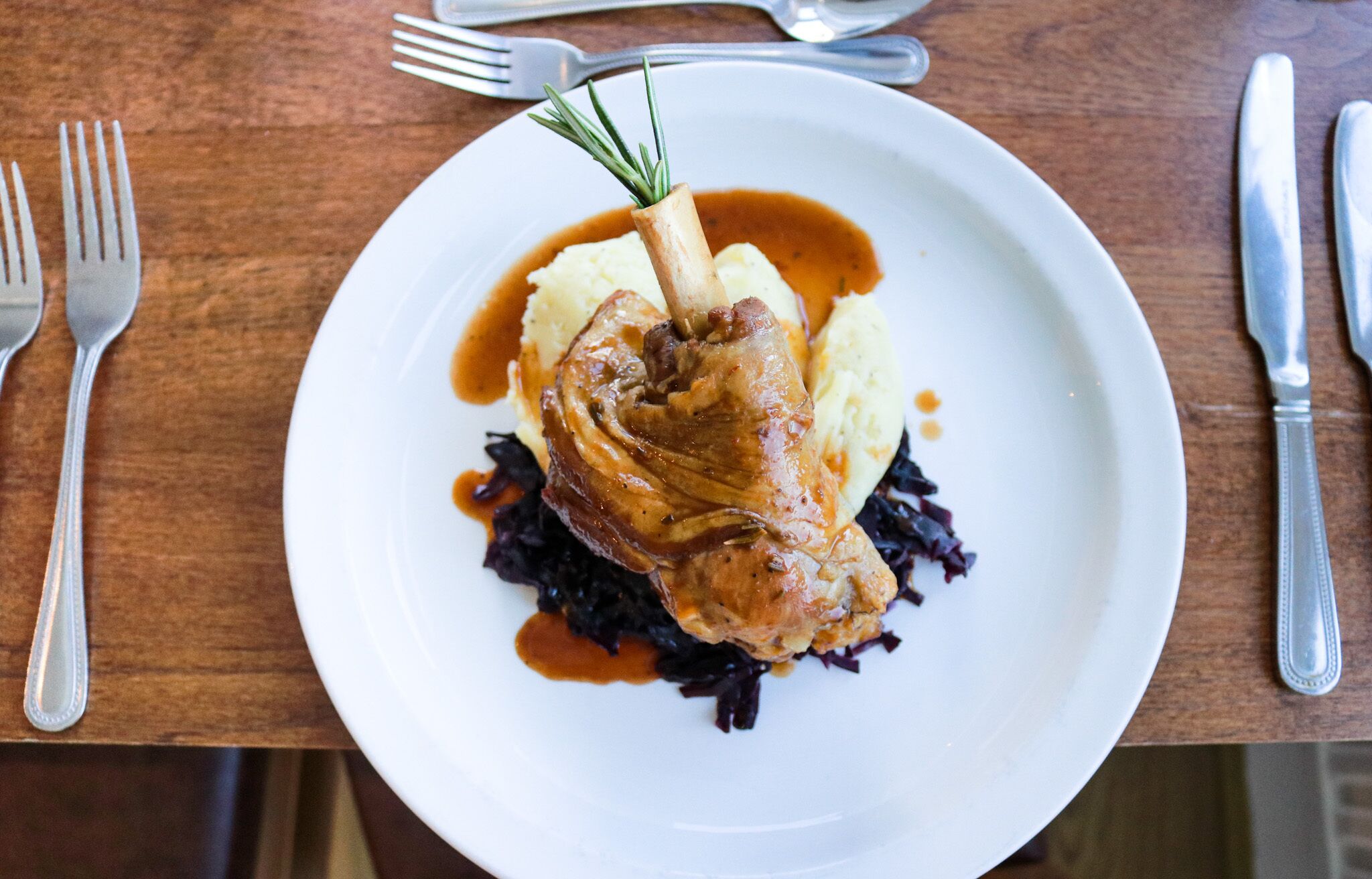 A plate with a roasted leg of lamb, mashed potatoes, and red cabbage, served on a wooden table with cutlery.