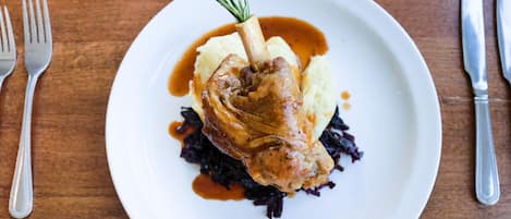 A plate with a roasted leg of lamb, mashed potatoes, and red cabbage, served on a wooden table with cutlery.